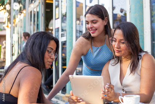 Three young women are sitting on the terrace of a restaurant, using a tablet to organize their week