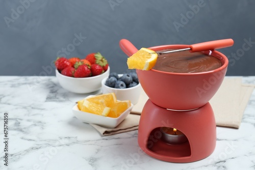 Fondue pot with melted chocolate, fresh orange, different berries and fork on white marble table. Space for text