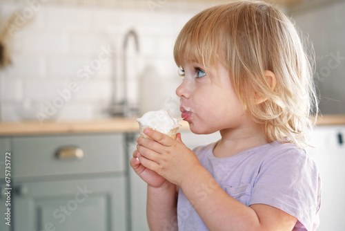 Baby girl enjoying ice cream. Pretty little toddler eating an ice-cream indoors, at home. Dining room background. Small child eats plombir and cream messy on her mouth. Cute kid with tasty sweet food.