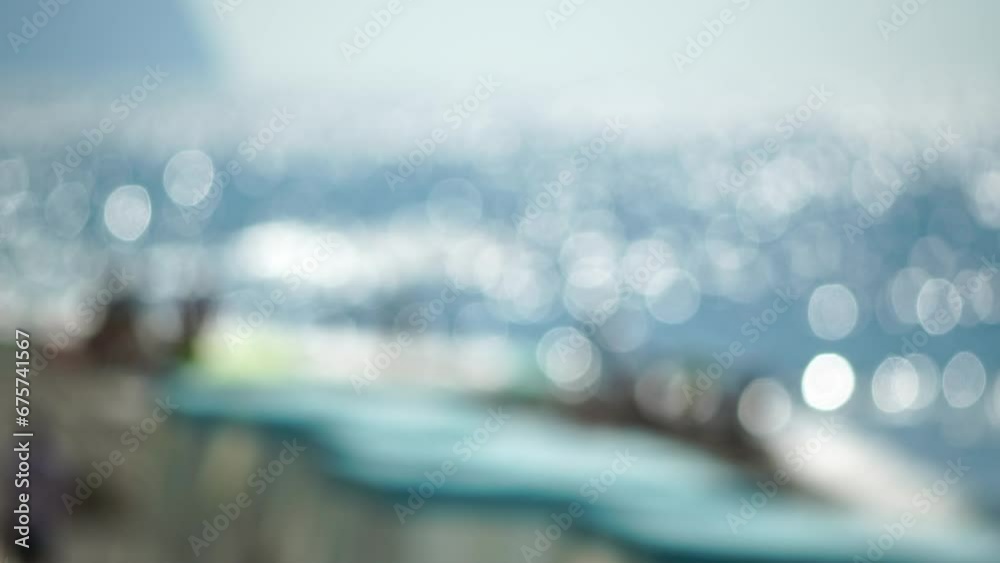Defocused sea ocean beach of hotel or resort with Lounge bed on hot summer day. Abstract bokeh background of blurred resting and swimming people on beach at blue sea. Holiday, vacation and recreation.