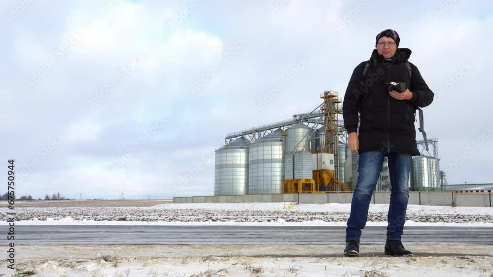 Photographer in the near silver silo on agro manufacturing plant for processing drying cleaning and storage of agricultural products. Grain silo elevator in winter in a field covered with snow Photographer in the near silver silo on agro manufacturing plant for processing drying cleaning and storage of agricultural products. Grain silo elevator in winter in a field covered with snow
