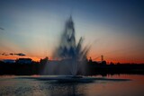 a water fountain spews water into the sky near a city at sunset