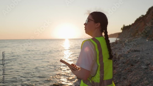 Side view of young Caucasian woman wearing eyeglasses and vest using tablet and walking on beach. Concept of environmental pollution and ecology research