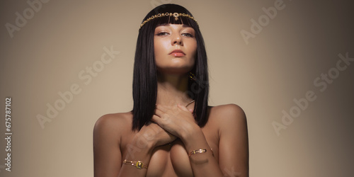 Beauty fashion portrait of naked woman queen cleopatra style covering her breasts by hands, looking at camera, posing bare with perfect make-up and golden jewelry 