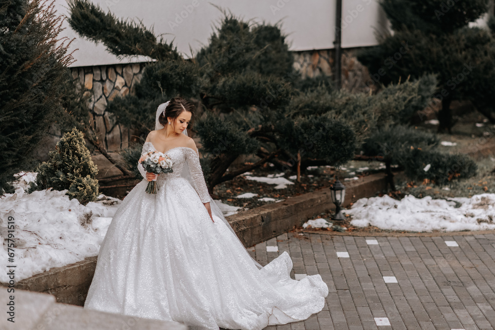 Fototapeta premium The bride in a white dress with a train and a long veil poses in nature, holding a bouquet. Walk. Winter wedding in nature.