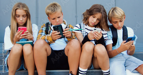 Group of teenage sad friends holding phones and browsing sitting on stairs near school building after lerning day. Happy positive teens friends texting on cellphones gadgets and talking. photo