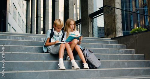 Cheerful high school students sitting on stairs street outdoors near campus talking smiling before classes. Caucasian boy and girl writing in their copybooks having an outdoor together after lessons