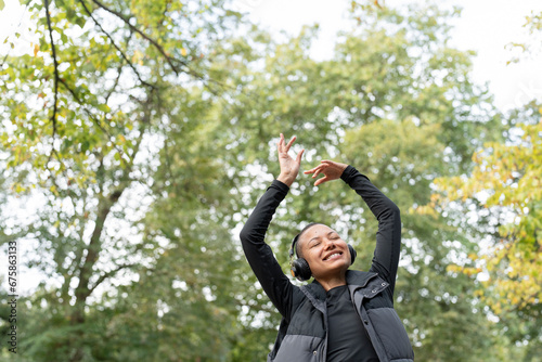 Portrait of smiling woman with headphones raising arms in park