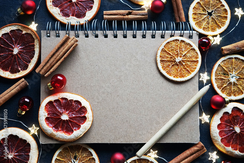 Notepad with pen next to dry fruit slices, Christmas tree decorations, cinnamon sticks on a dark background