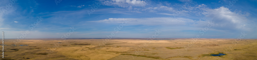 Panoramic aerial view of a vast dry prairie landscape under a blue sky with white clouds. A dirt road cuts across the fields and a small pond reflects the blue sky. One very lonely tree sits in the ce Panoramic aerial view of a vast dry prairie landscape under a blue sky with white clouds. A dirt road cuts across the fields and a small pond reflects the blue sky. One very lonely tree sits in the ce