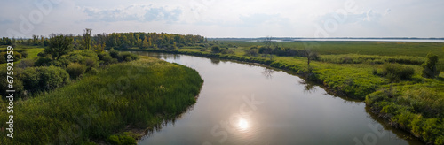Panoramic view of a curving river that cuts through marshy land and is reflecting the sun. The sky has a thin overcast with scattered clouds. 
