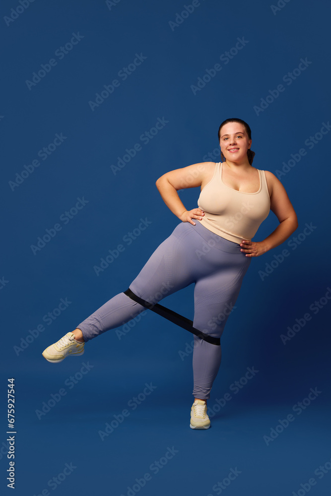 Leg swings. Young overweigh woman training with fitness elastic band against blue studio background. Concept of sport, body-positivity, weight loss, body and health care. Copy space for ad Leg swings. Young overweigh woman training with fitness elastic band against blue studio background. Concept of sport, body-positivity, weight loss, body and health care. Copy space for ad