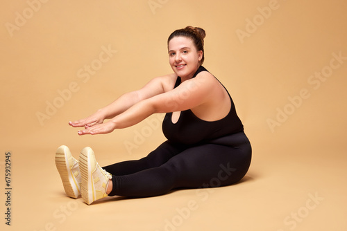 Young woman with fat, overweigh body in black sportswear sitting and doing stretching exercises against beige studio background. Concept of sport, body-positivity, weight loss, body and health care.