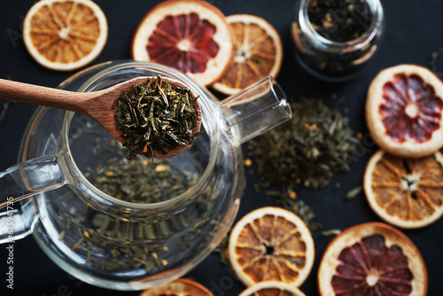 Loose leaf tea in a wooden spoon on the background of a glass teapot with tea and dry citrus slices