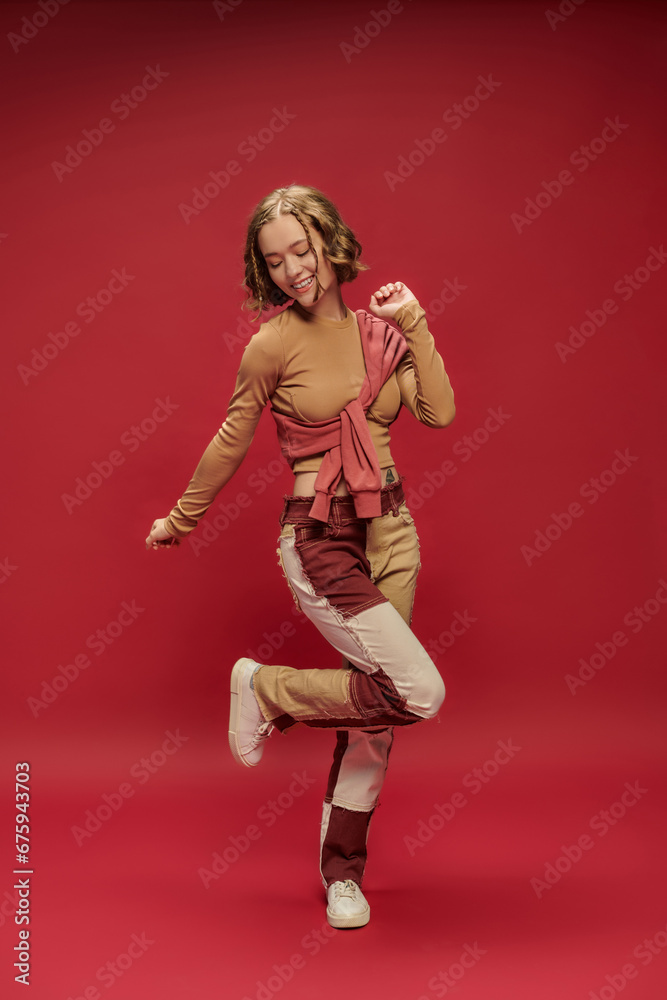 excited young woman in patchwork pants posing in jumper over cropped long sleeve on red backdrop excited young woman in patchwork pants posing in jumper over cropped long sleeve on red backdrop