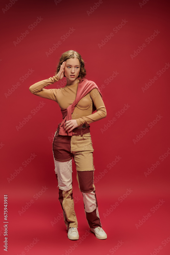 young trendy woman in patchwork pants posing in tied jumper over long sleeve on red backdrop young trendy woman in patchwork pants posing in tied jumper over long sleeve on red backdrop
