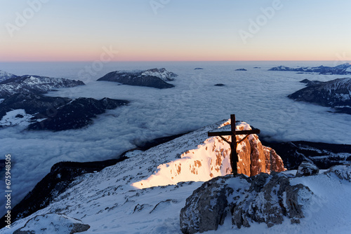 Sonnenaufgang am Watzmann Hockeck in den Berchtesgadener Alpen im Winter mit Blick auf das Gipfelkreuz und das Wolkenmeer über Berchtesgaden.