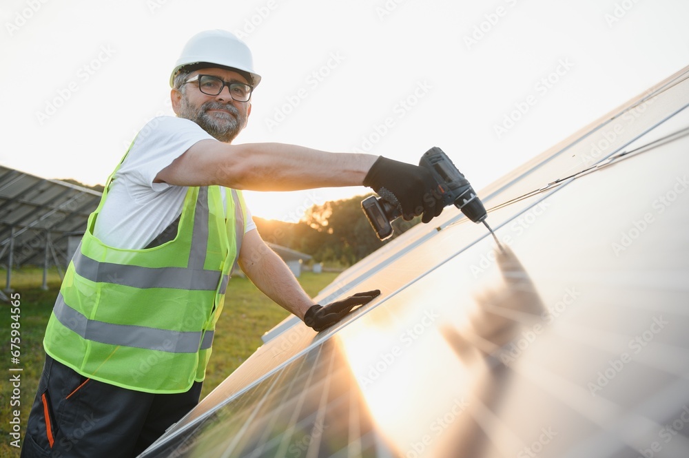 © Serhii - Portrait of smiling confident engineer technician with electrical screwdriver, standing in front of unfinished high exterior solar panel photo voltaic system