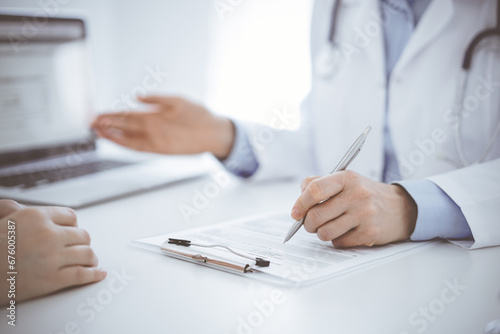 Doctor and patient sitting opposite each other at the desk in clinic. The focus is on female physician's hands filling up the medication history record form or checklist, close up. Medicine concept