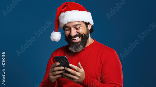 A man wearing a festive Santa hat is laughing with joy while looking at a smartphone in his hands against a solid background.