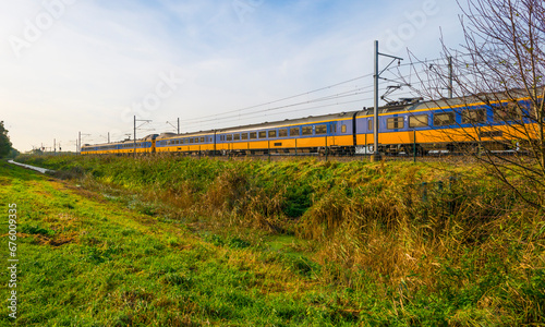 Train riding in a green field with reed in wetland beneath a blue cloudy sky in winter, Almere, Flevoland, The Netherlands, November 9, 2023