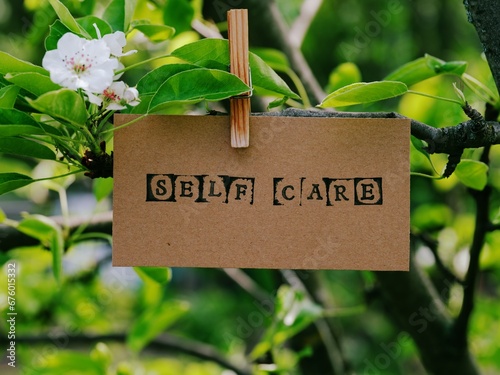 A piece of cardboard with the words Self Care on it hanging on a pear tree branch with blossoms and leaves using a wooden clothespin. © rosinka79