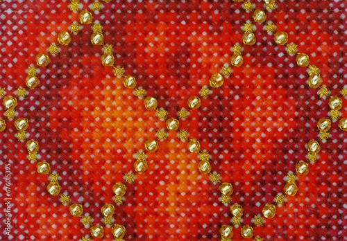 A close-up shot of a cross stitch maded with red and golden threads and gold beads.