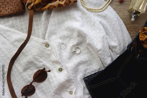 White blouse, denim skirt, brown shoes, vintage bag, leopard print scarf, pearl necklace, scrunchie, silver rings, perfume and lipstick on wood background. Retro, elegant outfit. Top view.