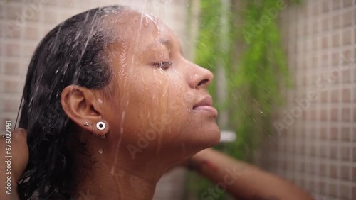 Young black woman taking relaxing shower at home. African American female wetting her hair and head with water enjoying self-care in bathroom. Concept of daily domestic personal hygiene in morning. photo