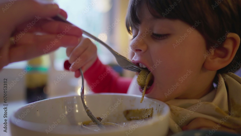 Mother feeding child spaghetti pasta food on bowl covered with napkin on collar. Little boy enjoys Italian food Mother feeding child spaghetti pasta food on bowl covered with napkin on collar. Little boy enjoys Italian food