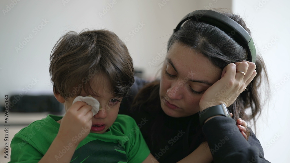 Tearful child holding cotton into hurt eye with patient but tired mother waiting for emergency service at hospital Tearful child holding cotton into hurt eye with patient but tired mother waiting for emergency service at hospital