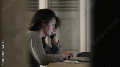 Woman browsing internet in front of laptop at home, candid authentic scene of person in front of computer screen late at night