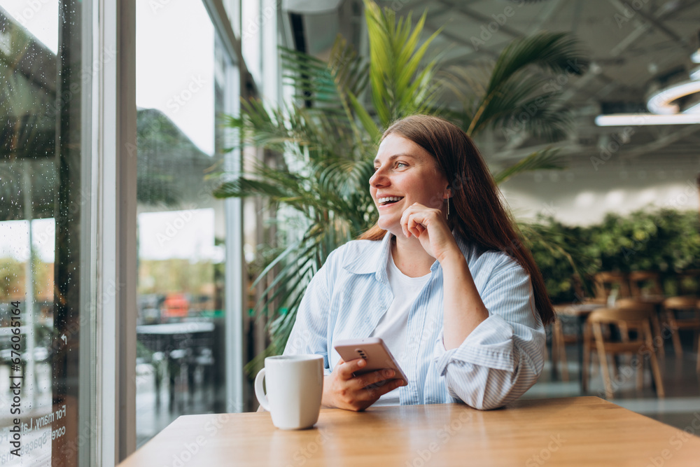 Young happy businesswoman smiling and using smart phone in a cafe. Person sitting at table and using smartphone indoors. Online education, order, working or shopping concept Young happy businesswoman smiling and using smart phone in a cafe. Person sitting at table and using smartphone indoors. Online education, order, working or shopping concept