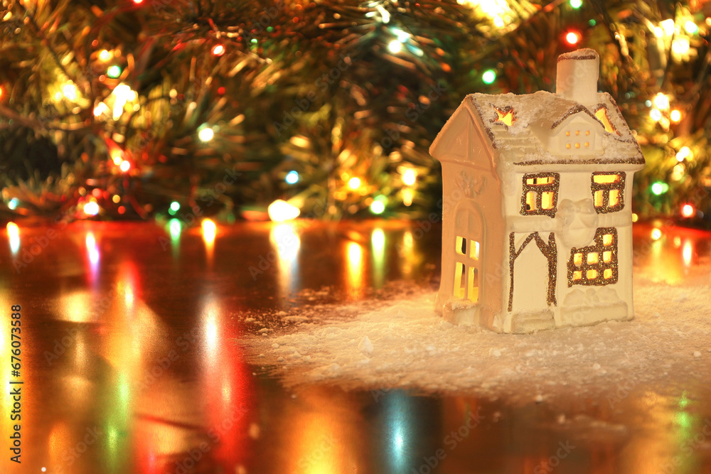Small ceramic white house candle holder lantern in decorative snow on table, Christmas tree with garland lights on background. Beautiful warm Xmas theme, New Year, winter time concept.