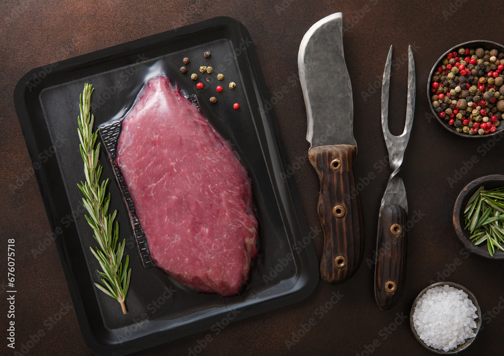 Beef raw fillet steak in vacuum tray with barbeque fork and knife on dark background with spices.