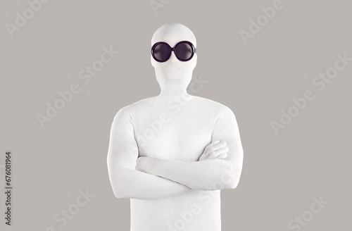Portrait of man disguised in bodysuit and sunglasses. Man wearing white skintight spandex body suit costume and black round glasses standing with crossed arms on grey color background photo