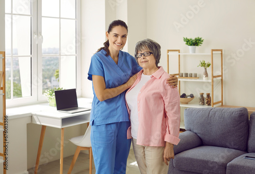 Young smiling brunette caregiver or nurse supporting and hugging senior woman patient at home. They are standing and looking at camera. Physiotherapist is helping old person, trust and care concept.
