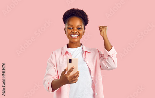 Overjoyed beautiful African American woman makes YES gesture with hand when sees notification on mobile phone about receipt of money or good news stands in pink studio. Ethnic girl portrait photo
