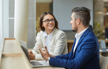 Two young smiling company employees man and woman sitting with laptop having discussion, talking about new job projects or startups. Business people working together in modern office.