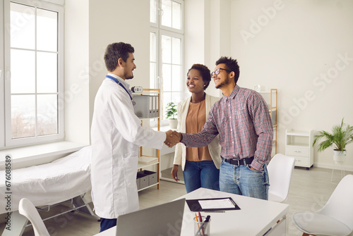 Smiling young multiracial couple handshake greeting with male doctor at consultation in clinic. Happy ethnic man and woman spouses shake hand make deal with GP in hospital. Healthcare concept.