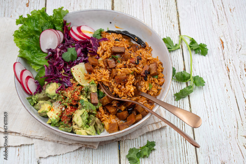 Mexican vegan bowl consisting of red ancho rice, avocado, red cabbage and seitan, greens, radishes. White bowl, wooden table. photo