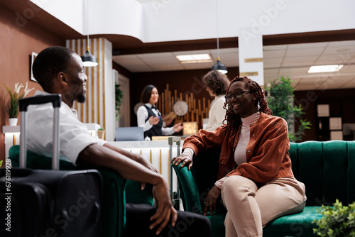Young couple waiting to do check in at hotel reception, relaxing on couch in luxury lounge area. African american people sitting in modern resort lobby before filling in registration forms.