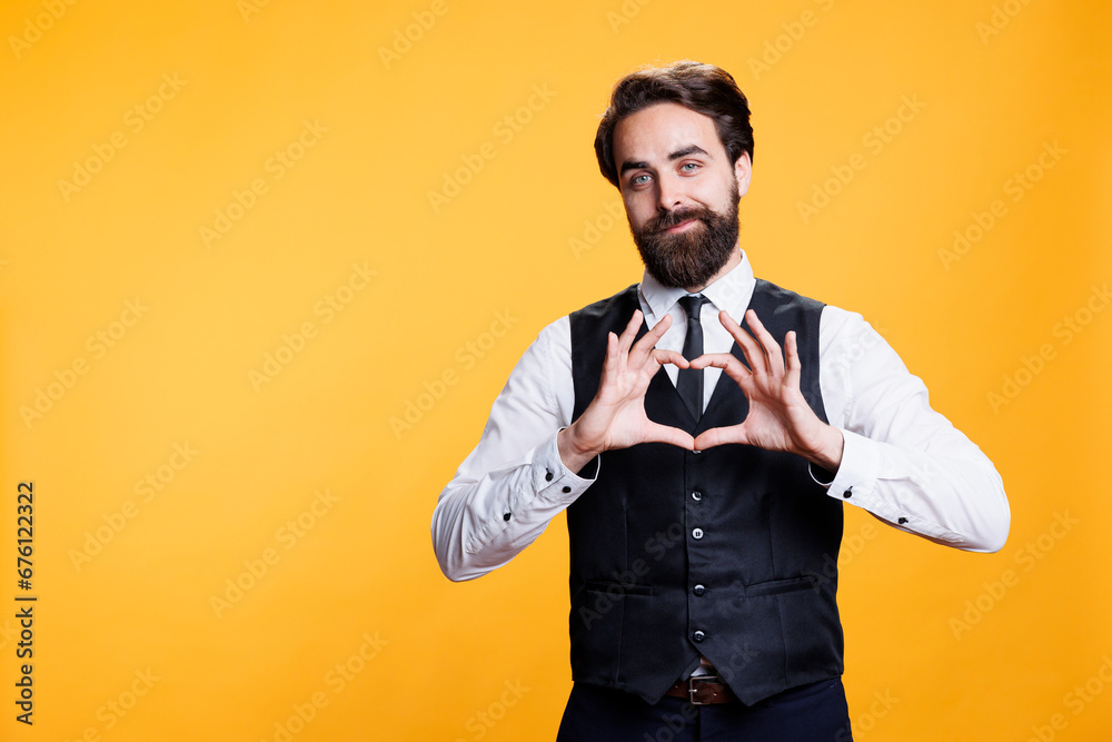 Butler presents heart shaped sign in studio, working at restaurant and showing true emotions on camera. Young adult acting romantic and expressing love with symbol, sincere feelings. - obrazy, fototapety, plakaty 