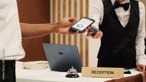 Receptionist using POS payment terminal to validate card purchase of room services and additional amenities, close up. African american guest using smartphone banking app during check in process