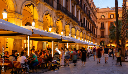 Citizens and tourists stroll and relax in cafe in light of streetlight on Royal square in Barcelona. Atmosphere of summer evening in Gothic quarter