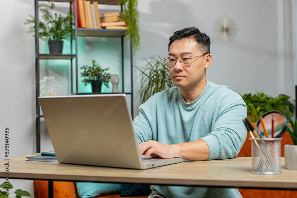 Confident concentrated Asian man working remotely on a project on laptop computer, thinking, typing on keyboard at home apartment. Freelancer guy communicates online chat sits on couch at table desk Confident concentrated Asian man working remotely on a project on laptop computer, thinking, typing on keyboard at home apartment. Freelancer guy communicates online chat sits on couch at table desk