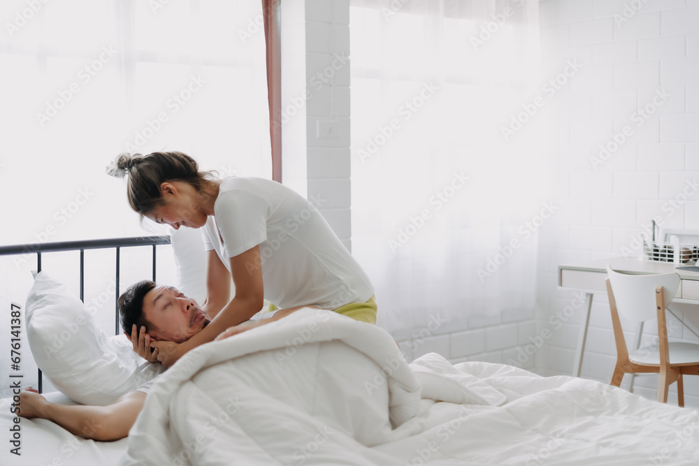 Funny asian couple wife try to wake lazy husband up in white bedroom. Funny asian couple wife try to wake lazy husband up in white bedroom.