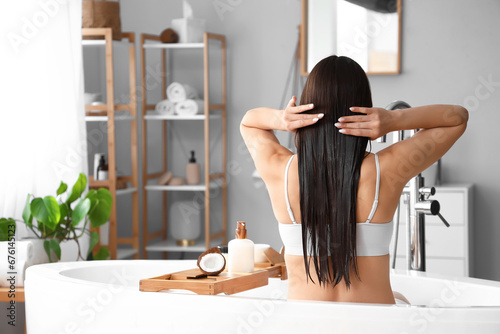 Beautiful young woman applying coconut oil onto her hair in bathroom, back view