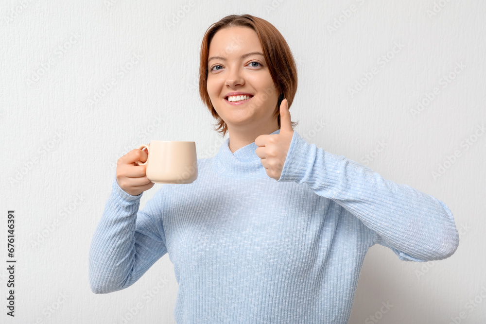 Young woman with cup of coffee showing thumb-up on white background