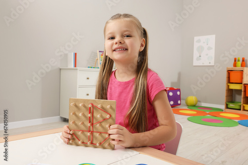 Motor skills development. Happy girl showing geoboard and rubber bands at white table in kindergarten photo